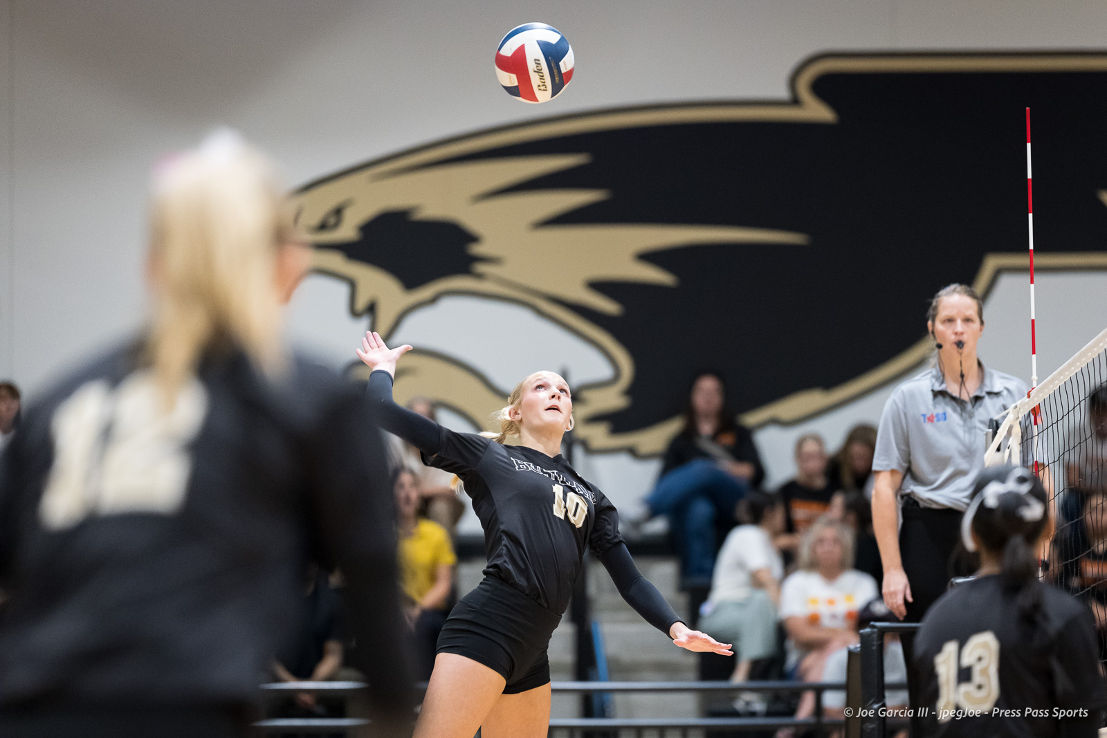 Volleyball player in black jersey (#10) serves the ball in a gym with a large team logo on the wall, referee visible at right and net in frame.