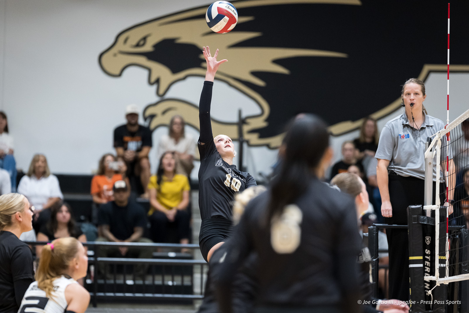 Volleyball player in black jersey (#10) leaps to strike the ball as the referee watches, with spectators in the gym behind her.