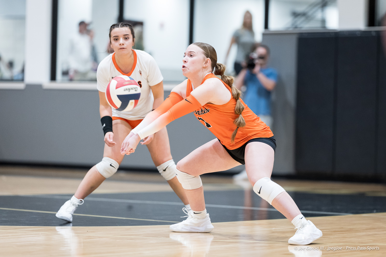 Two female volleyball players in orange and white uniforms dive for a ball on an indoor court, faces focused as they reach for the ball in play.