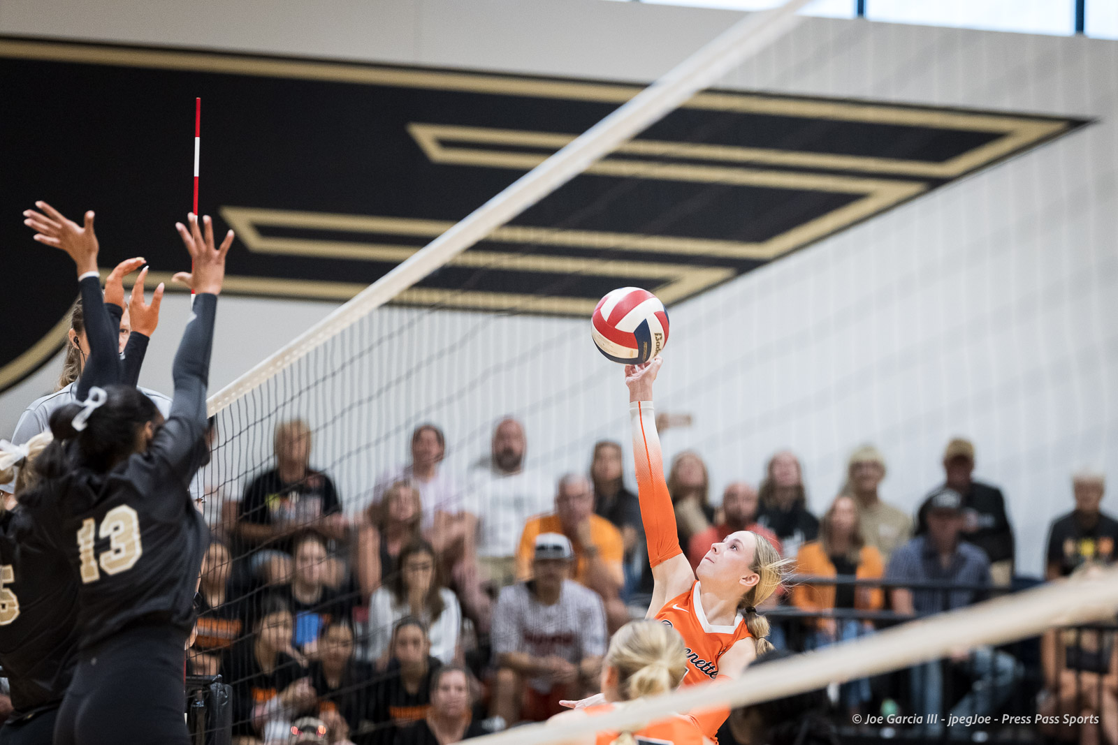 Orange-team player spikes the volleyball over the net while a defender in black jumps to block.