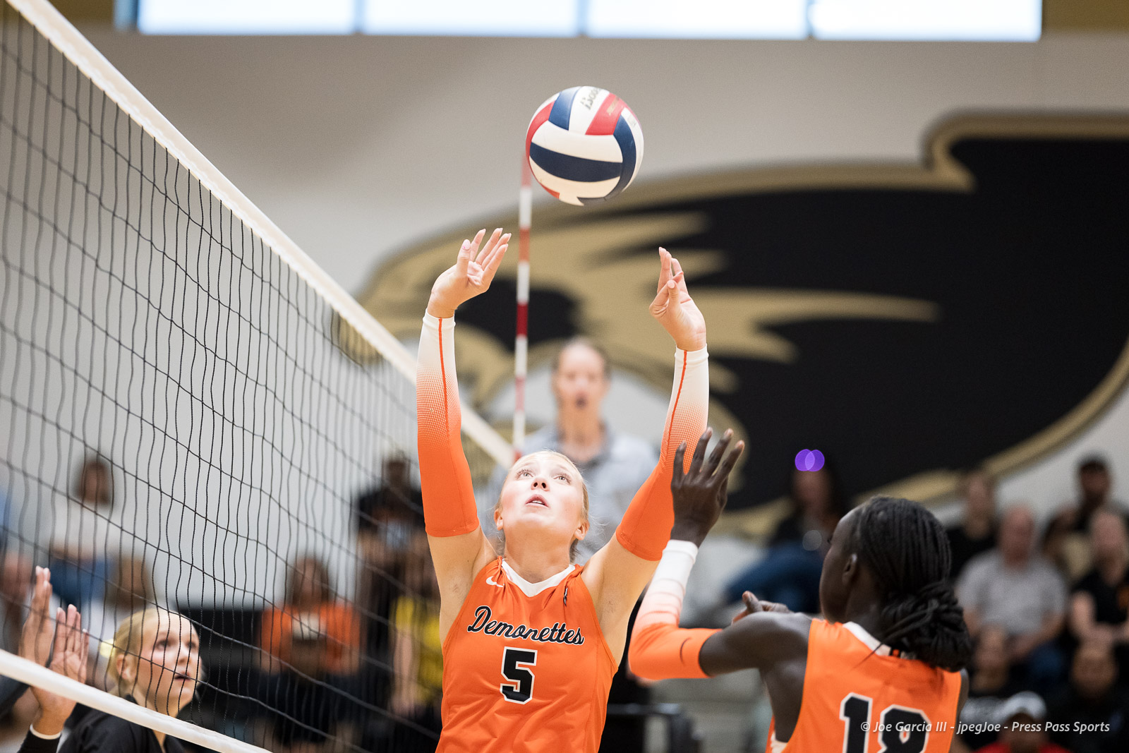 Female volleyball player in orange jersey (#5) reaches up at the net to block a ball, crowd watching in the background.
