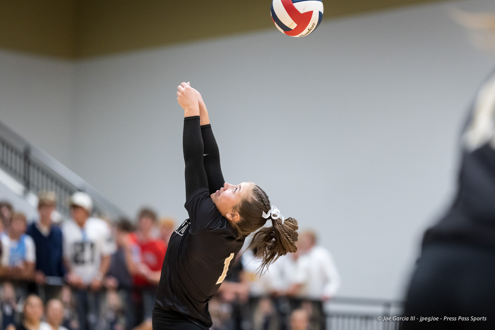 Female volleyball player in a black uniform with a white bow in her hair, arms extended upward to set a volleyball, with a blurred crowd watching in the background.