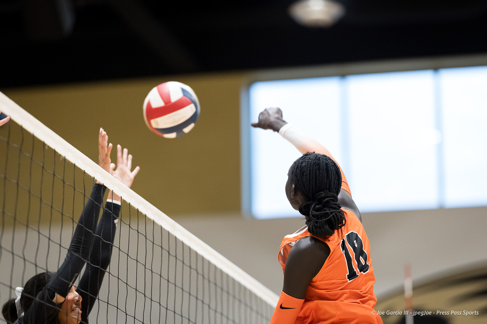 Volleyball player in orange jersey (#18) spikes the ball over the net as opponents reach up to block indoors.