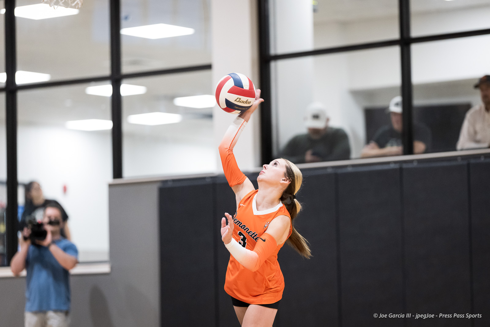 Female volleyball player in an orange uniform serves the ball indoors, with spectators and a photographer in the background.