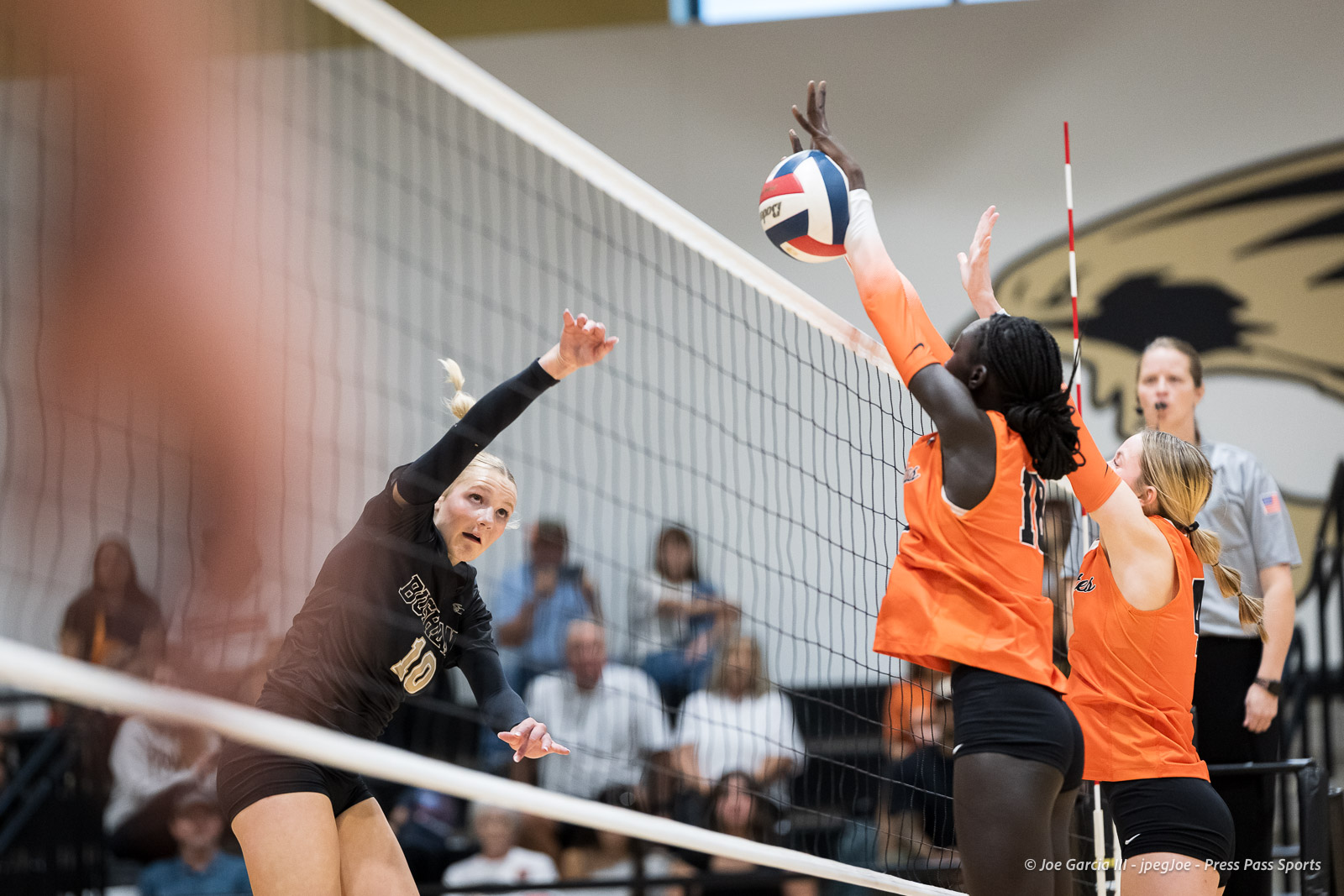 Left-side player in black spikes the ball toward the net as orange-jersey blockers leap to block it at the net indoors.