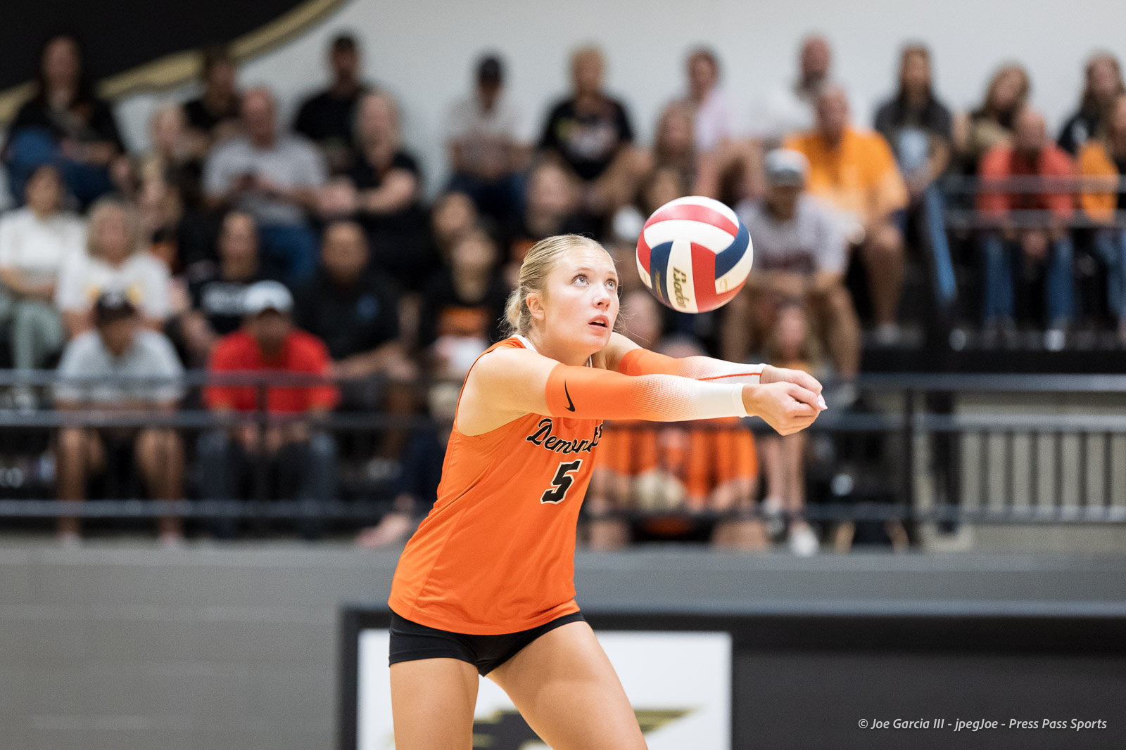 Female volleyball player in an orange jersey stretches to pass the ball during a match, with a blurred crowd in the stands behind her.