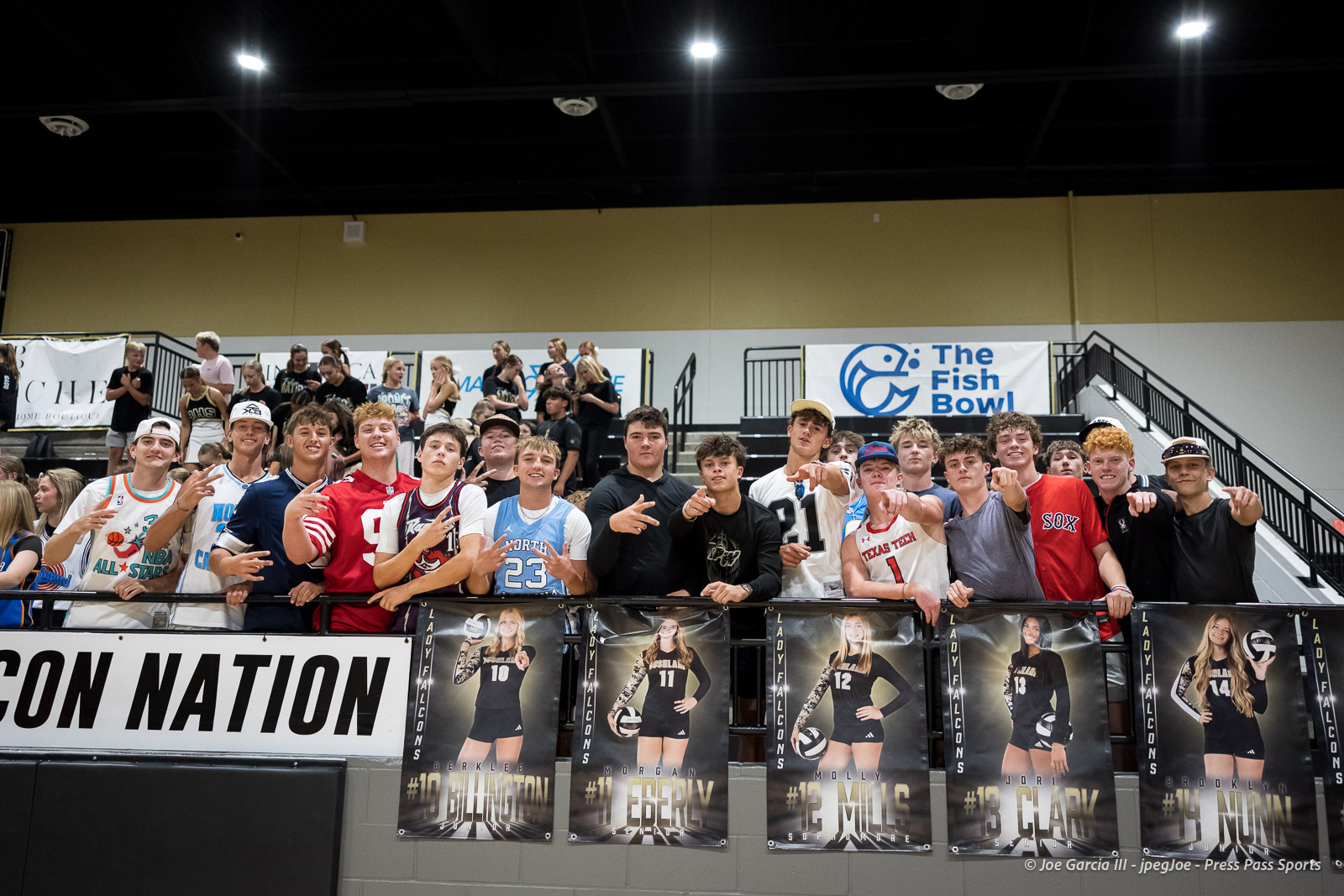 Group of young athletes and fans leaning on a railing in an indoor gym, with posters of players hanging below them.