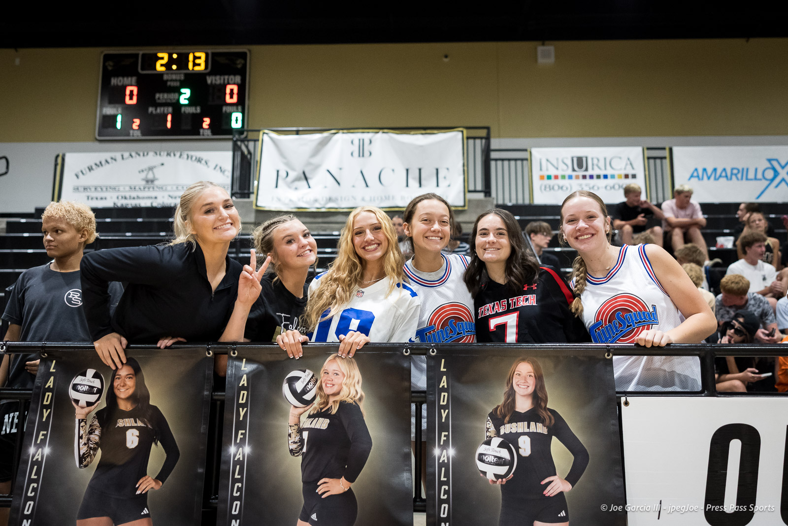 Six smiling female volleyball players lean over a railing in a gym, posing for the camera with banners in the background.