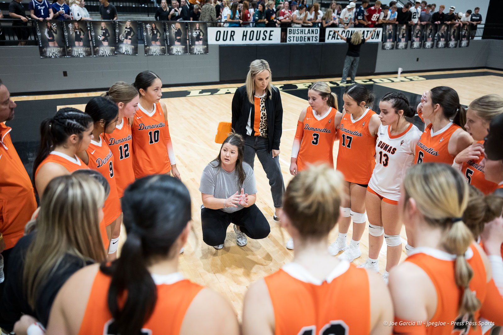 Girls' basketball team gathers in a circle around a kneeling coach during a timeout on a gymnasium floor.