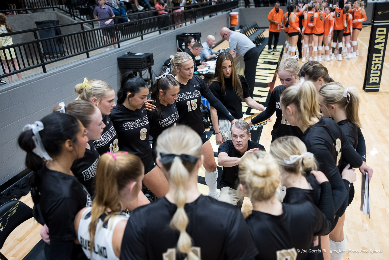Bushland girls' volleyball team huddling around their coach on the court during a timeout; players in black jerseys.