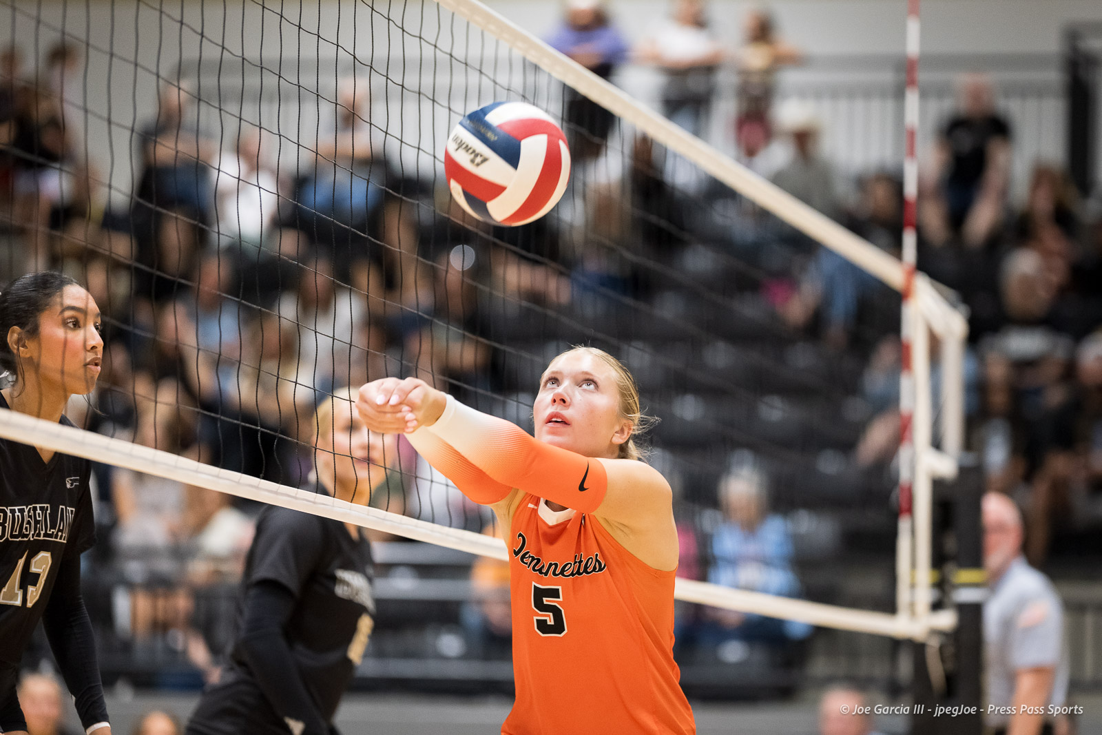 Orange-jersey volleyball player #5 reaches toward the ball at the net during a match, with a black-jersey opponent nearby and a blurred crowd in the background.