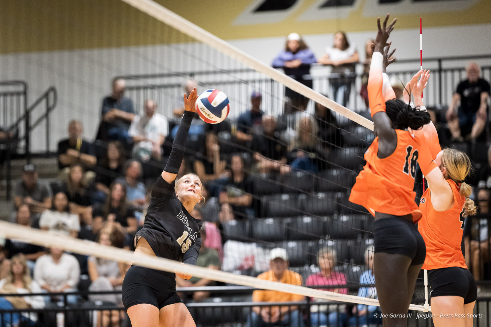 Black-uniform player spikes a volleyball past two orange-jersey blockers at the net, in a crowded gym.