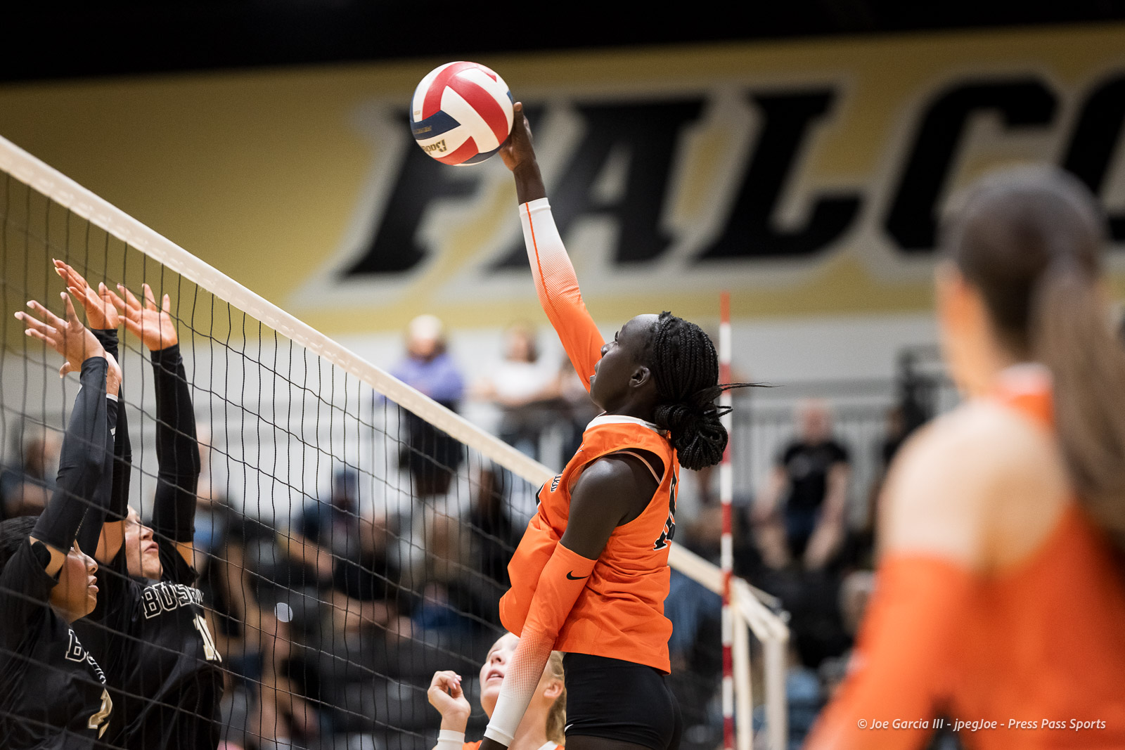 Volleyball player in orange spikes the ball over the net toward defenders in black jerseys.