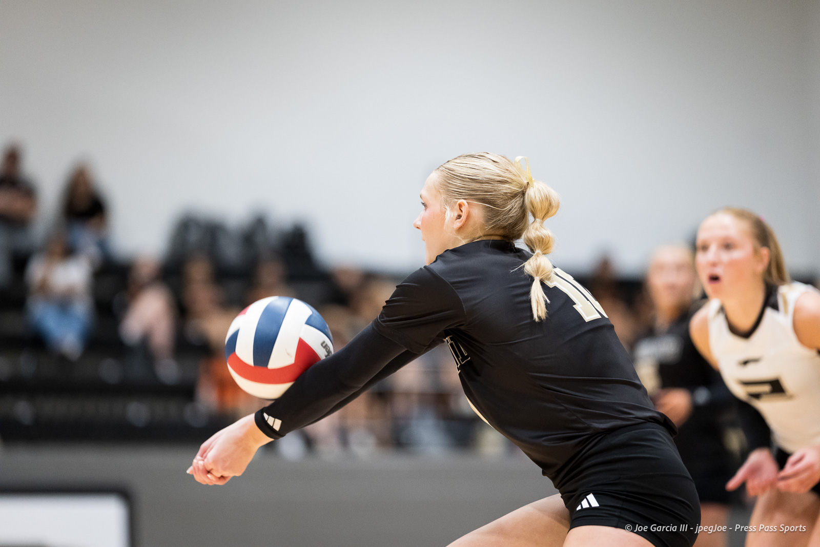 Volleyball player in a black uniform sends the ball with extended arms during a match, with a teammate and blurred crowd in the background.