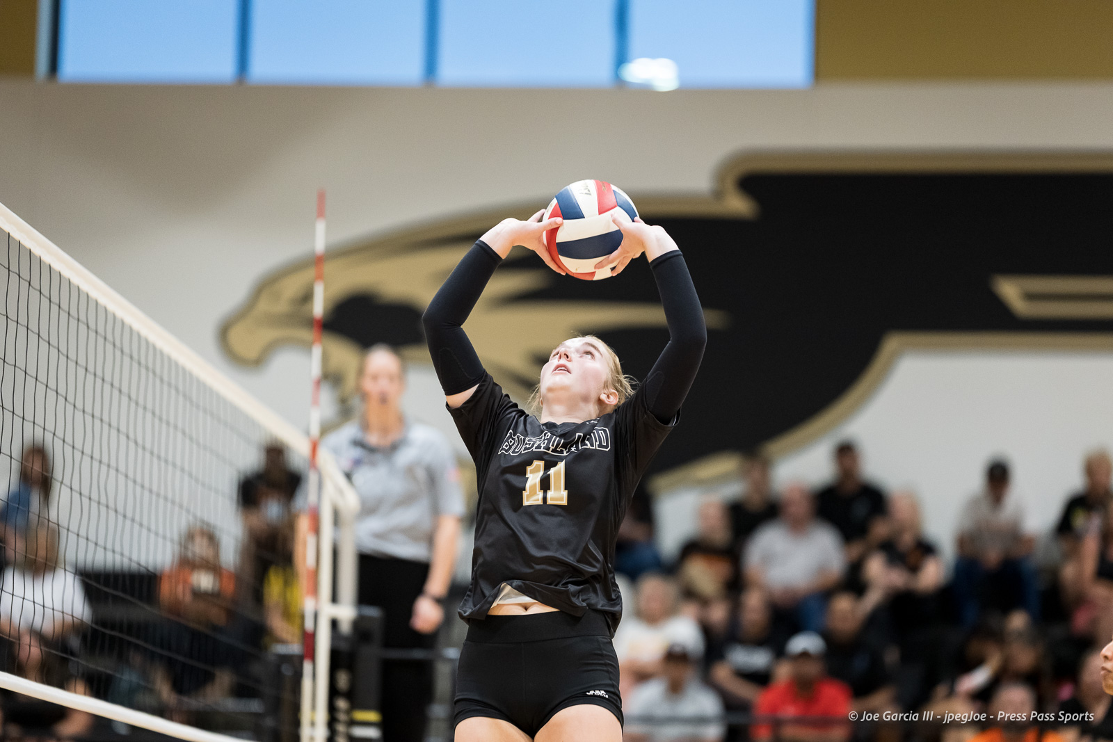 Female volleyball player with jersey number 11, jumping to set a ball over the net in a gym with spectators in the background.