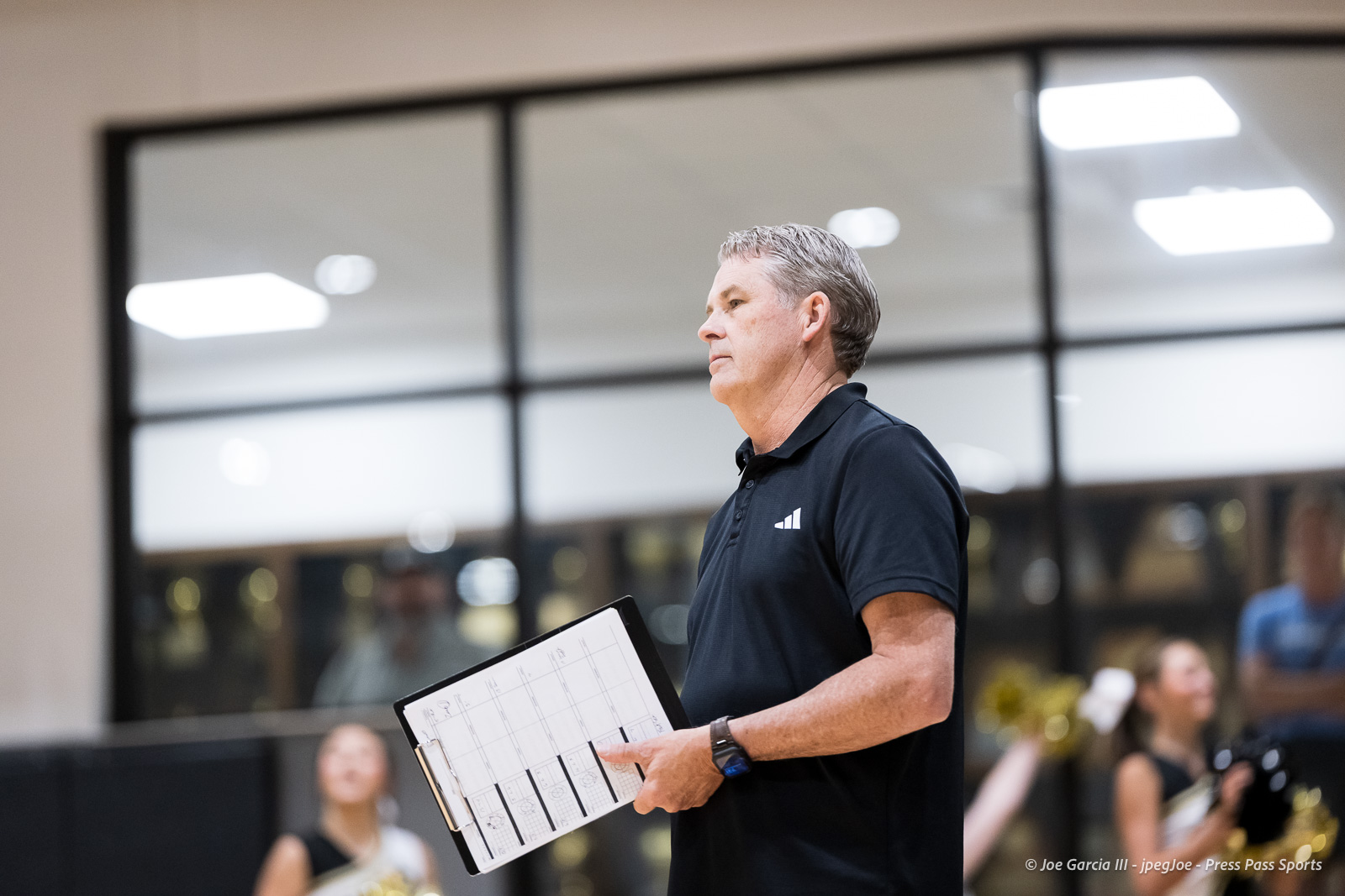 Midday scene of a man in a black polo speaking or presenting, holding a clipboard with a calendar/plan in a gym or rehearsal room.