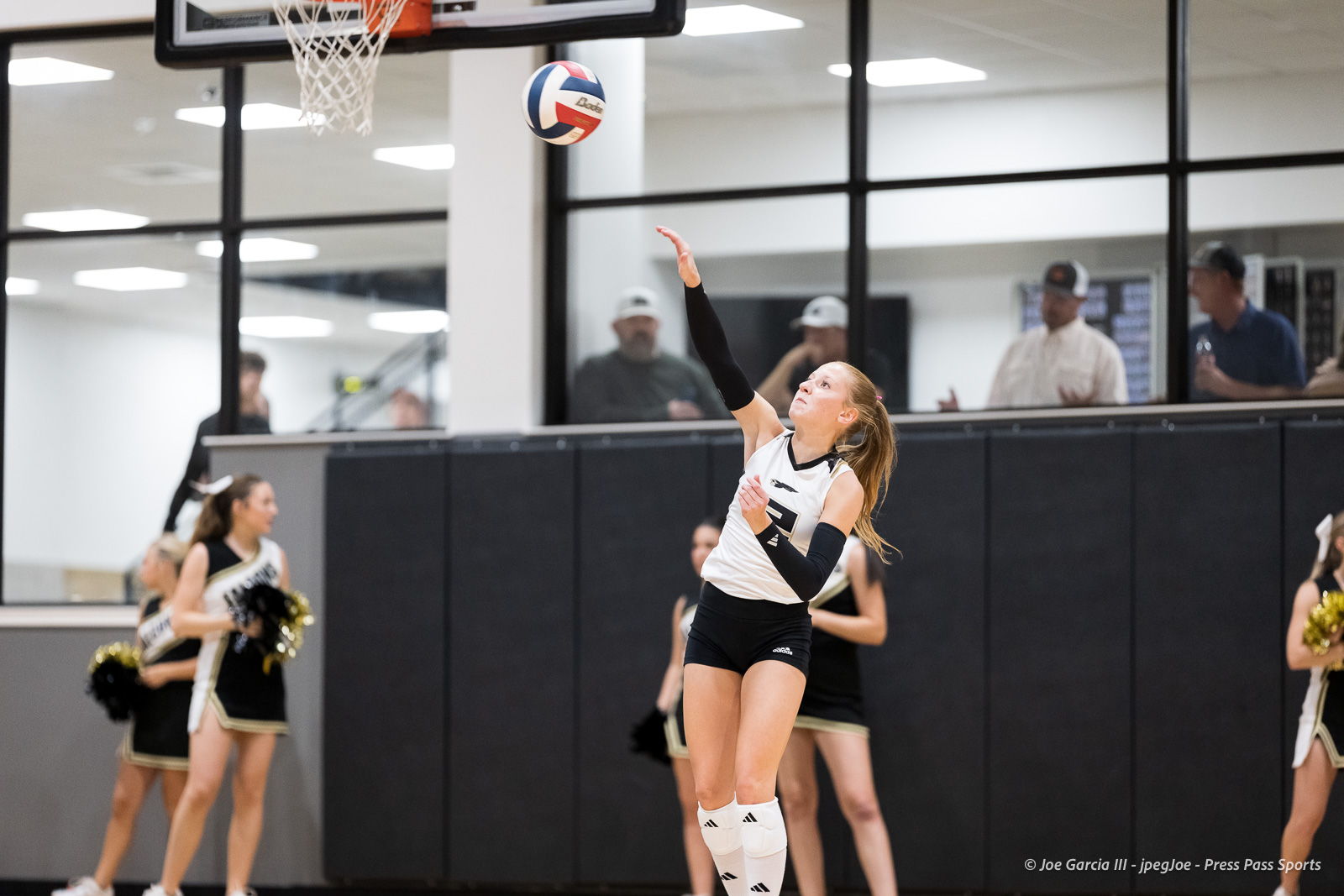 Female volleyball player in white and black uniform serves the ball in an indoor gym; cheerleaders and spectators watch from the background.
