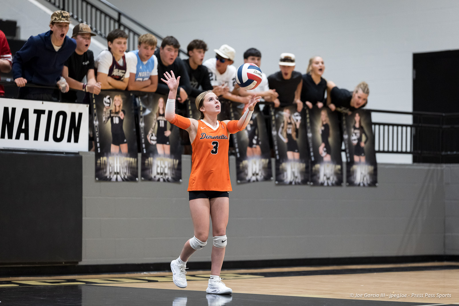 Volleyball player in orange jersey #3 serves the ball as a cheering crowd watches from the stands behind her.