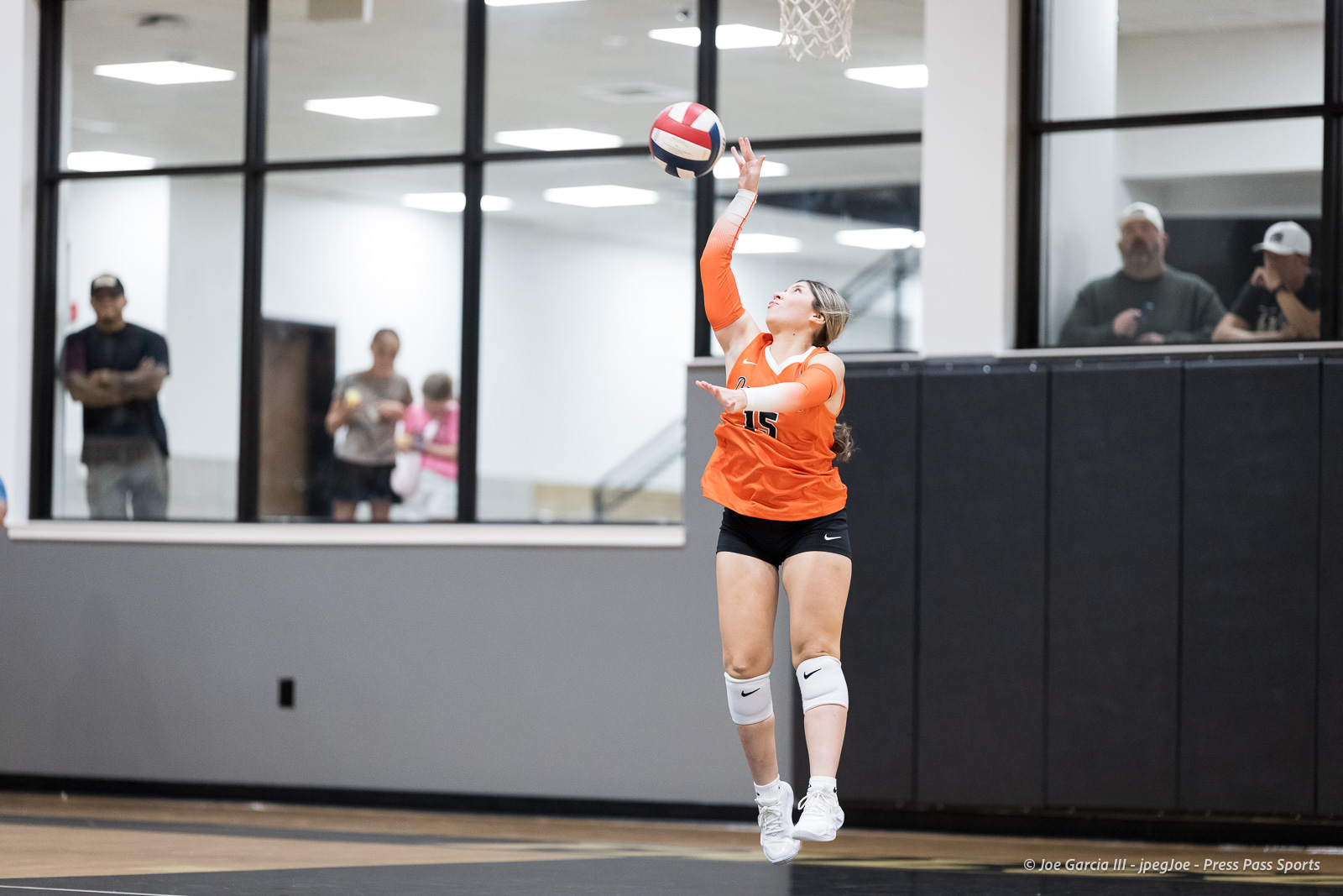 Female volleyball player in orange jersey jumps to hit a ball during a match inside a gymnasium, spectators in the background.
