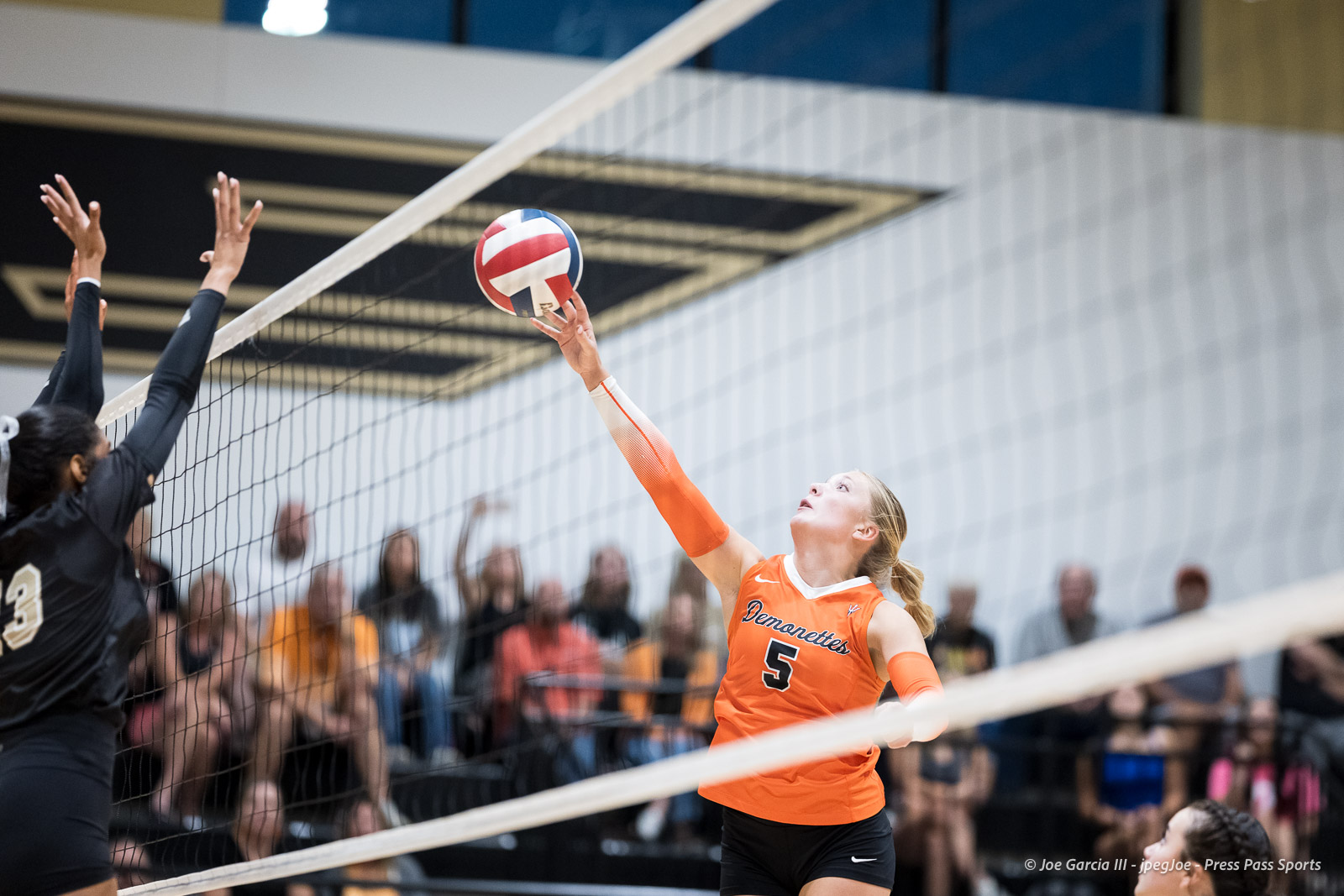 Orange-clad volleyball player spikes the ball over the net as a defender reaches up to block, with a cheering crowd in the background.
