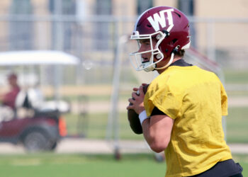 Football player in a yellow practice jersey holding a ball, wearing a maroon helmet on a sunny field.