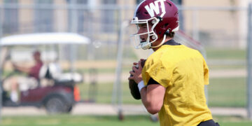 Football player in a yellow practice jersey holding a ball, wearing a maroon helmet on a sunny field.