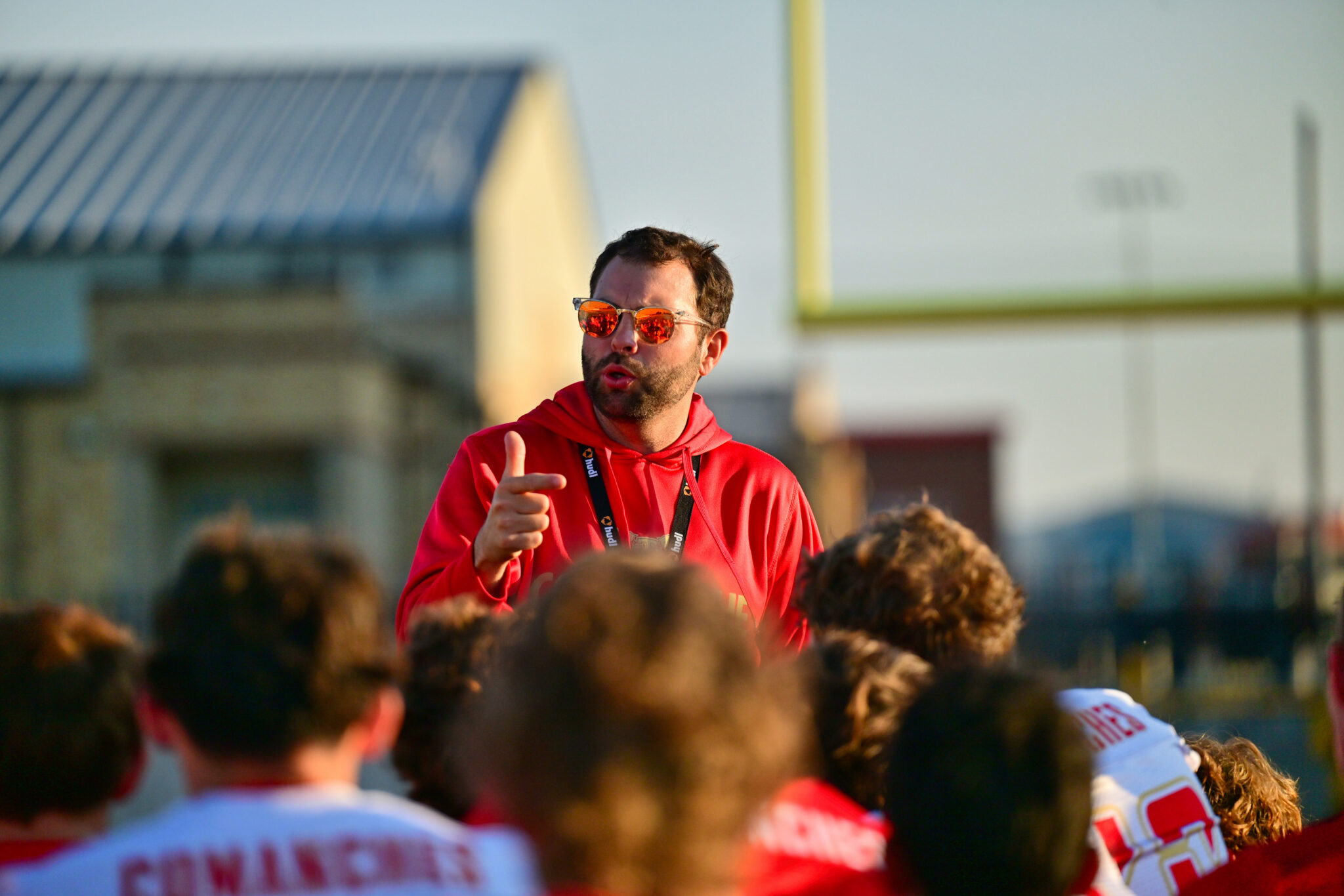 DSC_8691 | Press Pass Sports Man in a red hoodie with orange sunglasses addressing a crowd outdoors at a sports event.