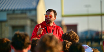 Man in a red hoodie with orange sunglasses addressing a crowd outdoors at a sports event.