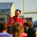 Man in a red hoodie with orange sunglasses addressing a crowd outdoors at a sports event.