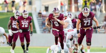 West Texas A&M quarterback #9 runs with the ball as teammates block near a sunny football field.