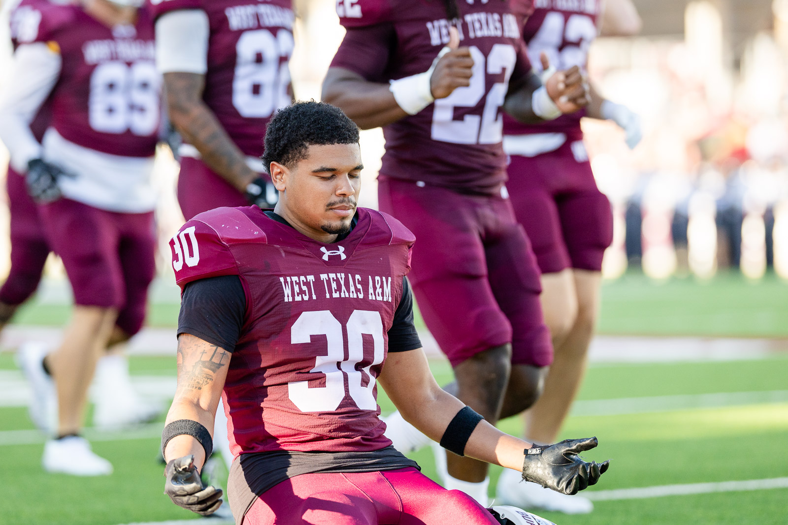 western-colorado-vs-wtamu-jpegJoe-pps-1 | Press Pass Sports West Texas A&M football player #30 sits on the field with eyes closed, surrounded by maroon-uniform teammates.