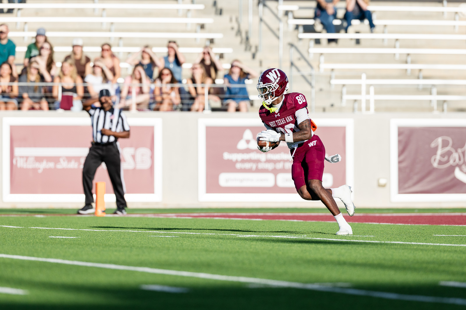 western-colorado-vs-wtamu-jpegJoe-pps-3 | Press Pass Sports Football player in a maroon uniform runs with the ball on a green turf field, crowd in stands in the background.