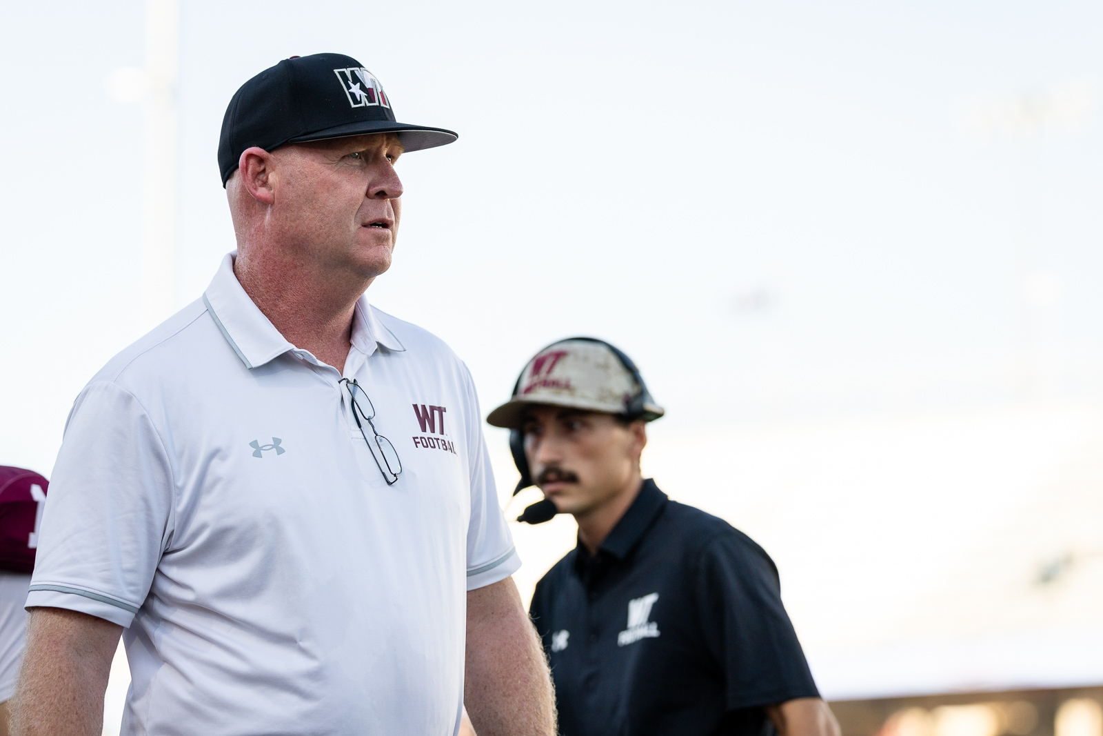western-colorado-vs-wtamu-jpegJoe-pps-4 | Press Pass Sports Football coach in a white polo with glasses hanging from the collar stands on the sideline, wearing a black cap with a logo; another staffer is blurred in the background.