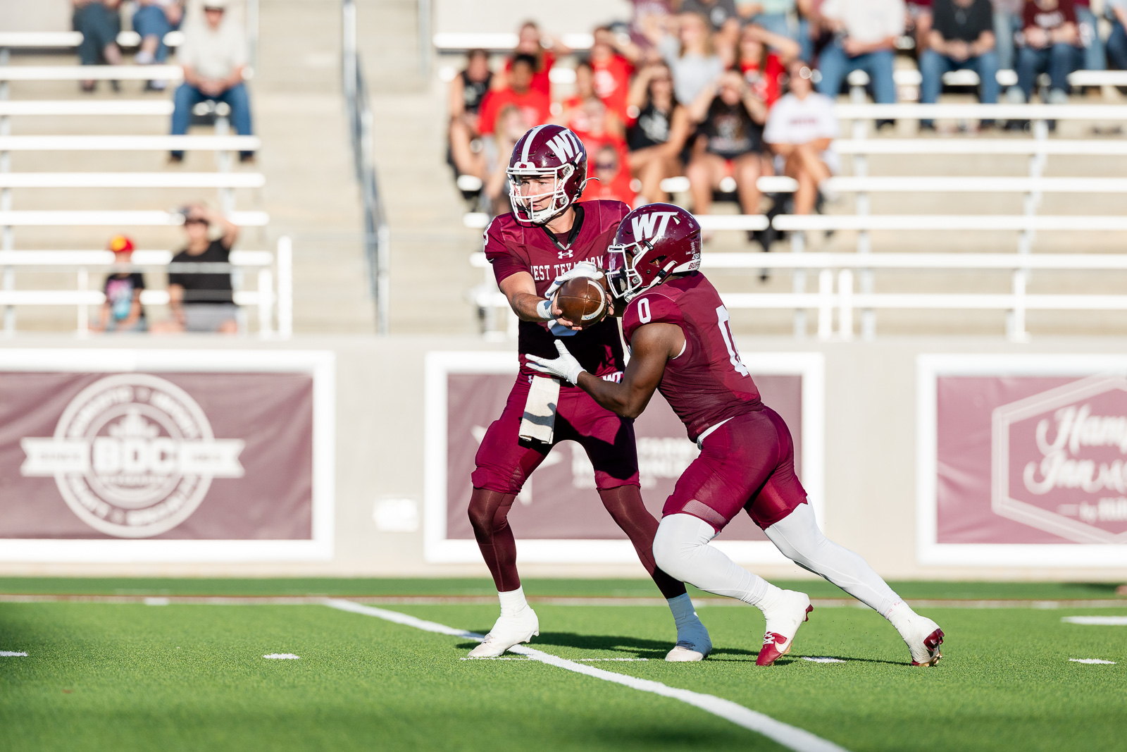 western-colorado-vs-wtamu-jpegJoe-pps-5 | Press Pass Sports Two college football players in maroon uniforms contest a loose football on a bright green field, with spectators in the stands watching nearby.