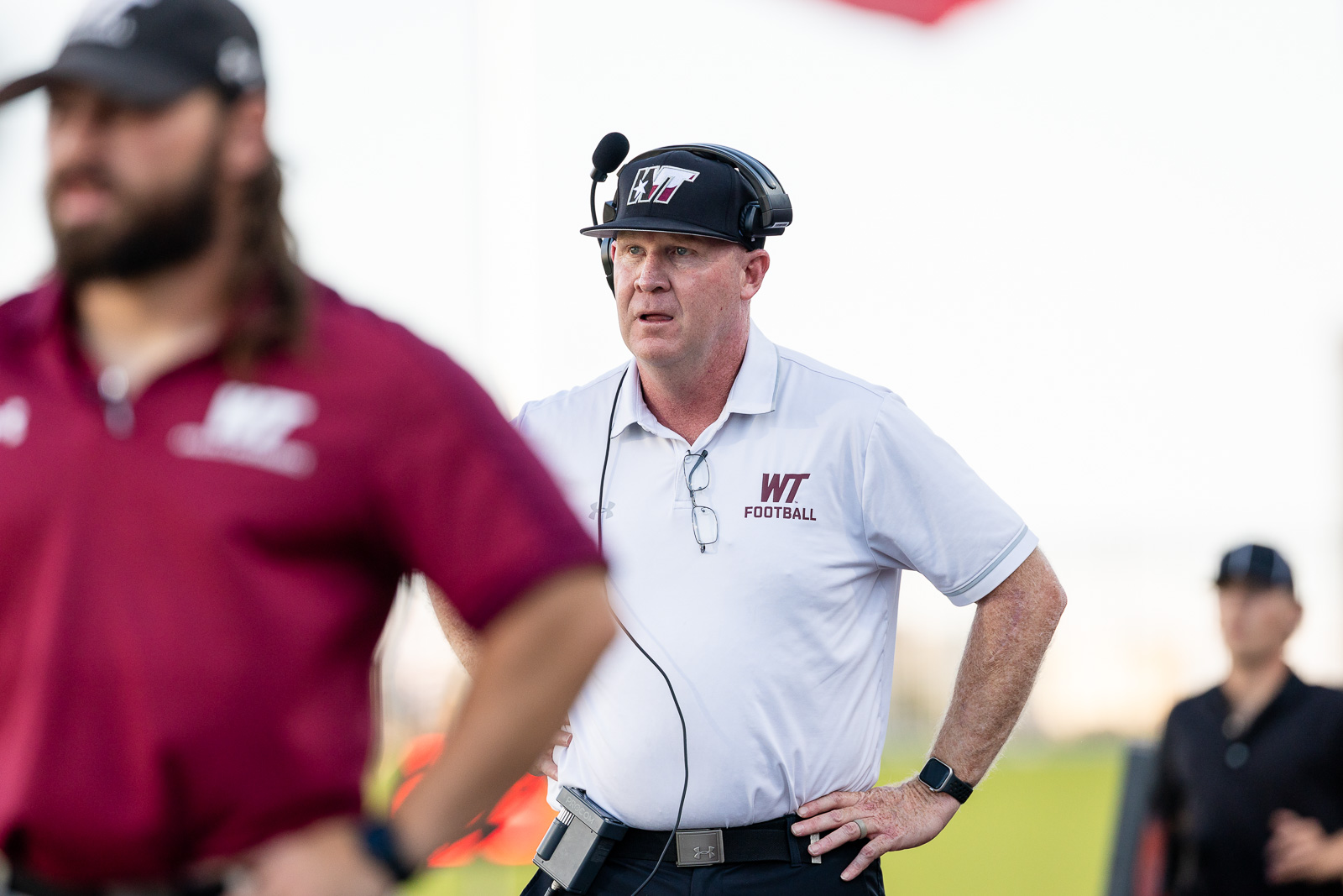 western-colorado-vs-wtamu-jpegJoe-pps-7 | Press Pass Sports Coaching staff member wearing a headset and WT Football polo stands on the sideline watching play, hands on hips.