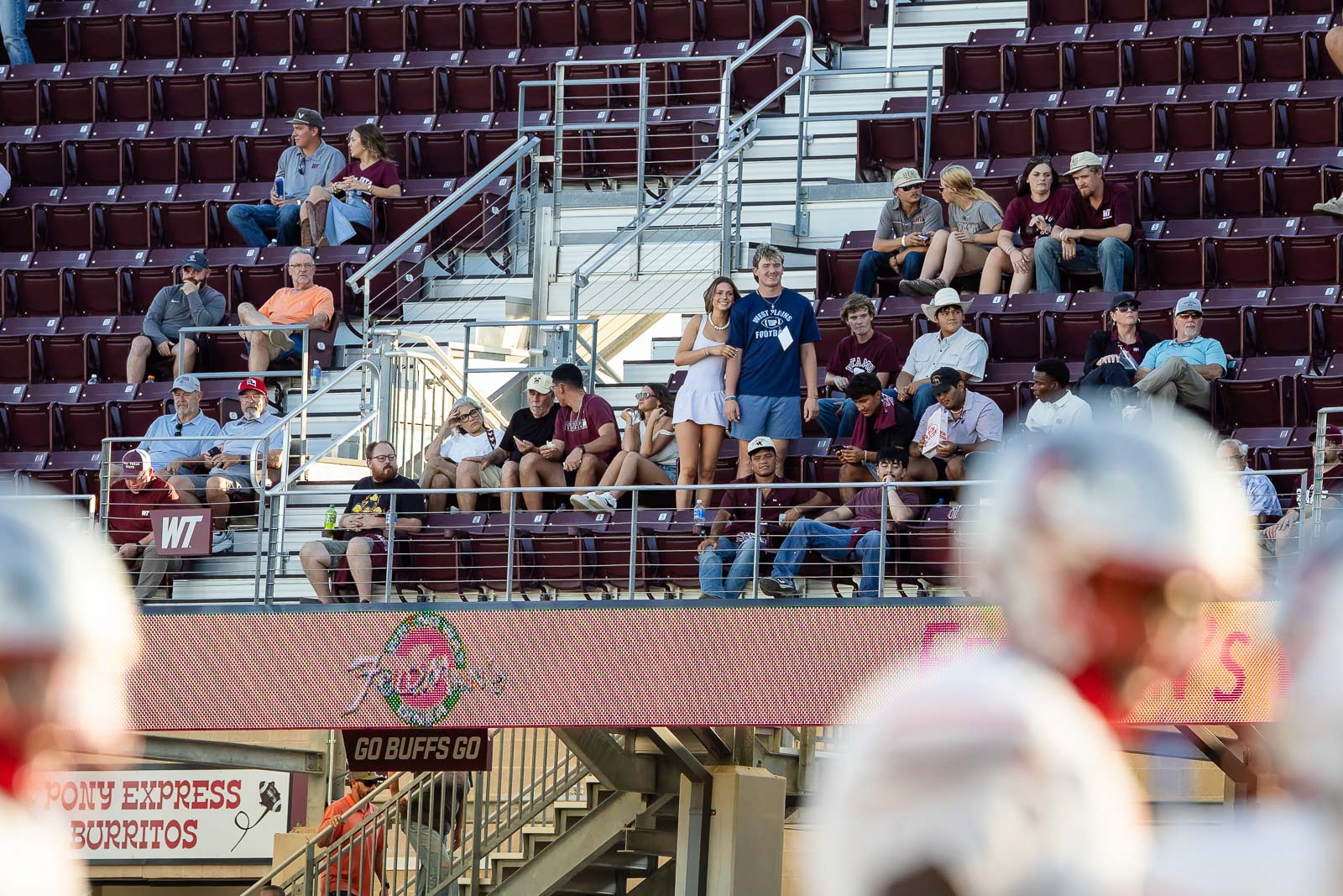 western-colorado-vs-wtamu-jpegJoe-pps-19 | Press Pass Sports Couple standing in the stands beside a football field crowd, other spectators around them and ad boards below.