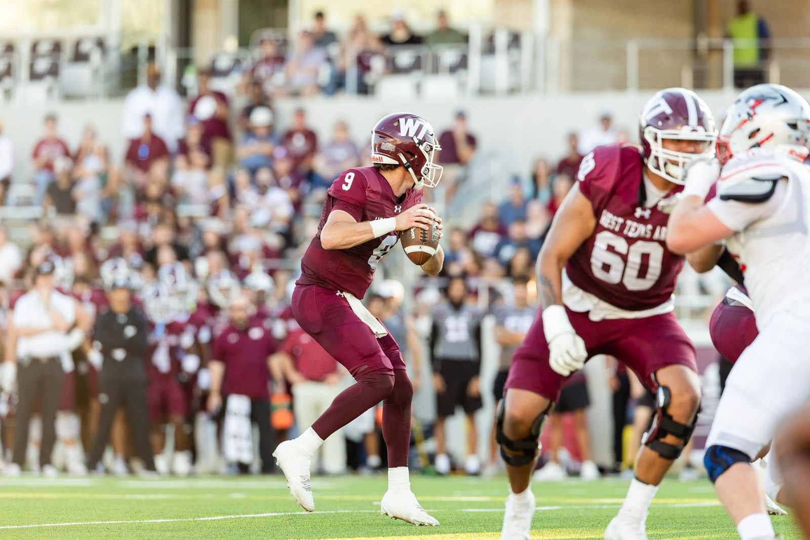 western-colorado-vs-wtamu-jpegJoe-pps-22 | Press Pass Sports Maroon-clad quarterback #9 drops back to pass during a college football game, with teammates, opponents, and a cheering crowd in the background.