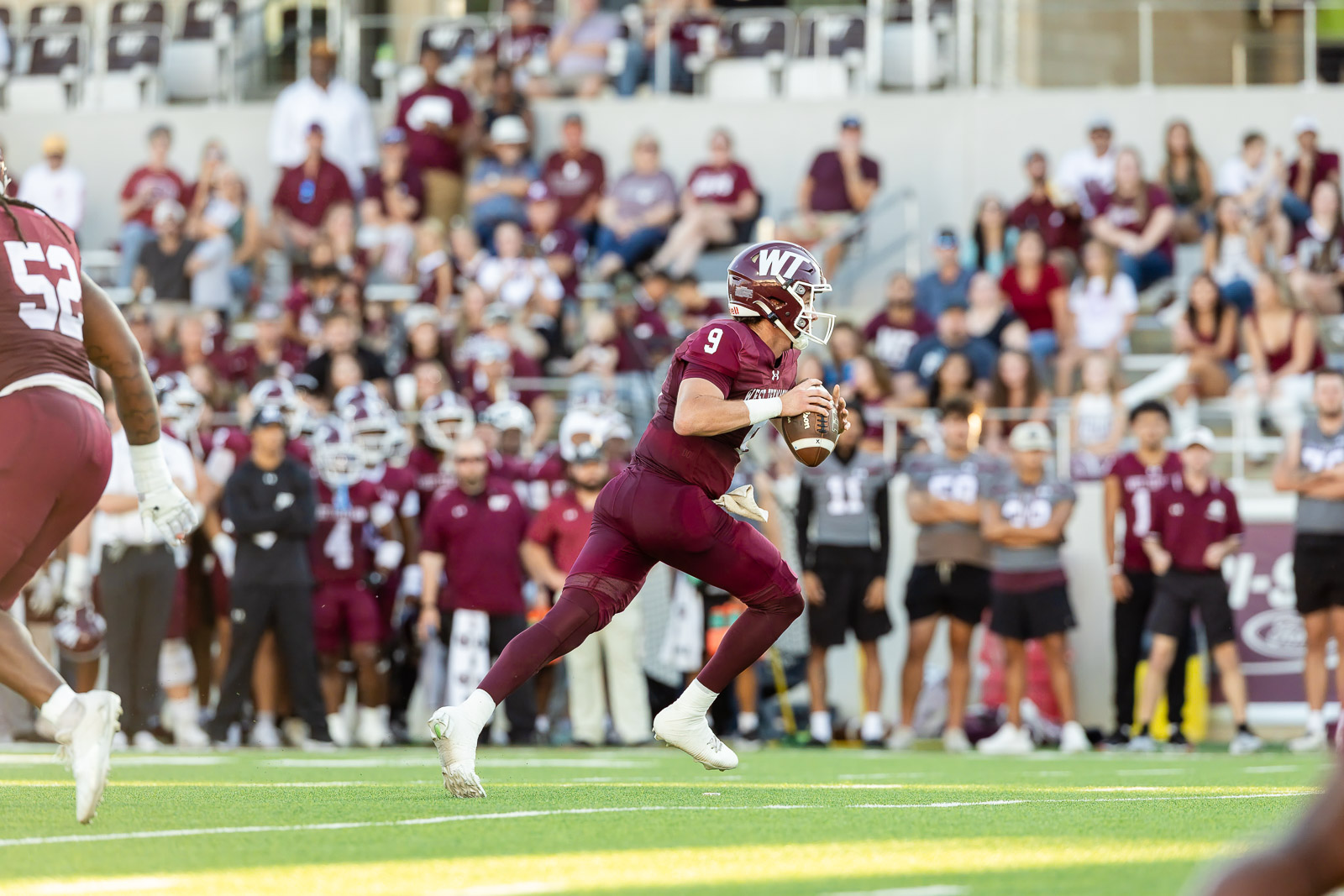 western-colorado-vs-wtamu-jpegJoe-pps-23 | Press Pass Sports Maroon-clad quarterback (#9) sprints with the ball during a college football game, crowd cheering in the stands behind him.