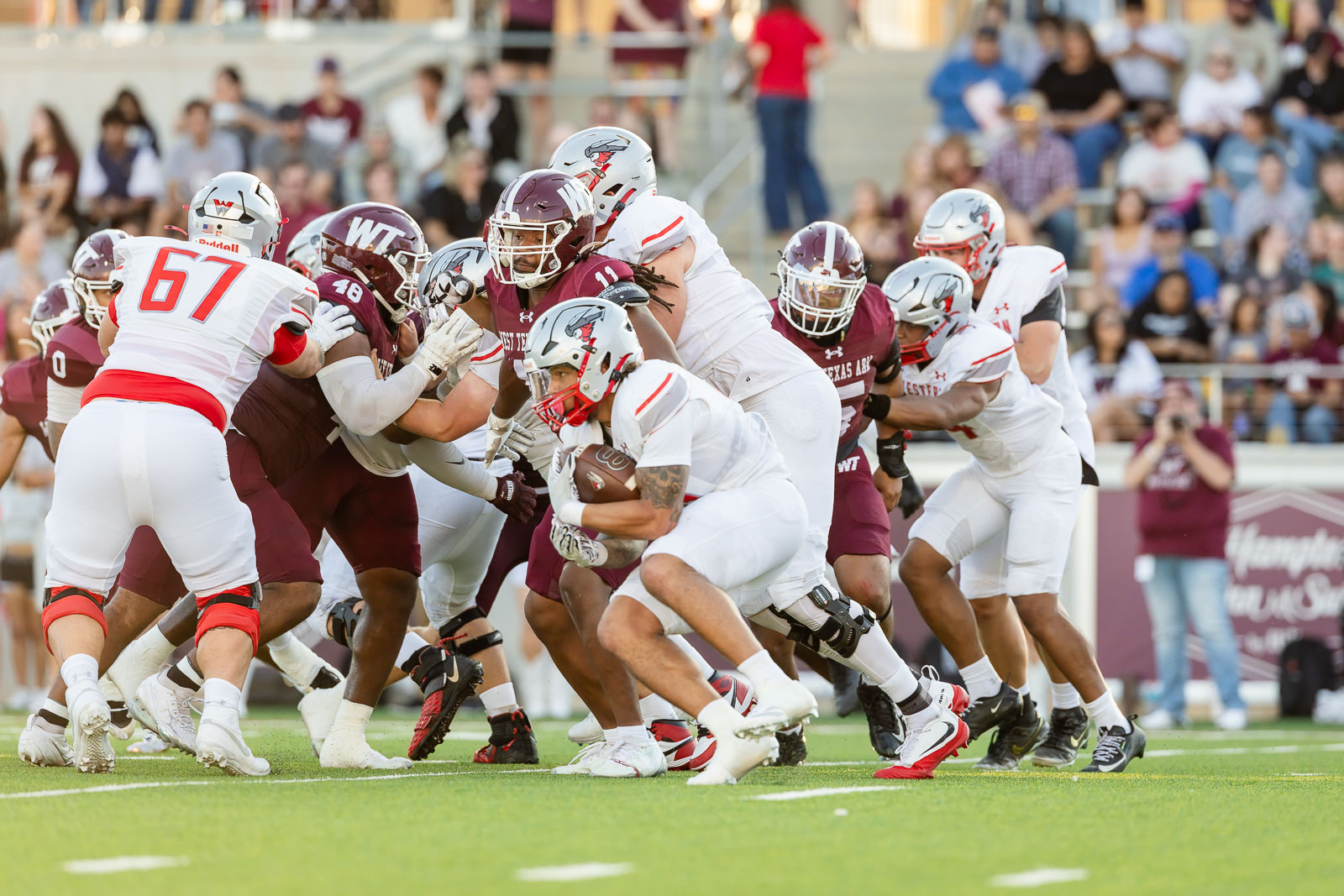 western-colorado-vs-wtamu-jpegJoe-pps-24 | Press Pass Sports Group of college football players in maroon and white collide at the line of scrimmage as a ball carrier in white advances guarded by blockers. Fans watch from the stands in the background.