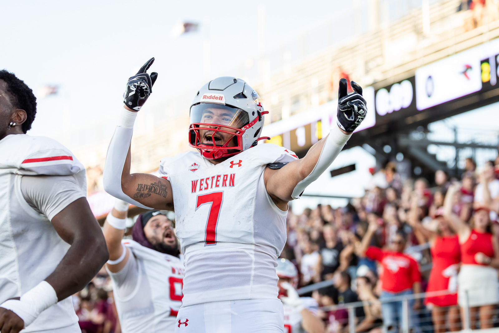 western-colorado-vs-wtamu-jpegJoe-pps-30 | Press Pass Sports Football player in white uniform with number 7 raises arms in celebration on the field; crowd in stands behind.