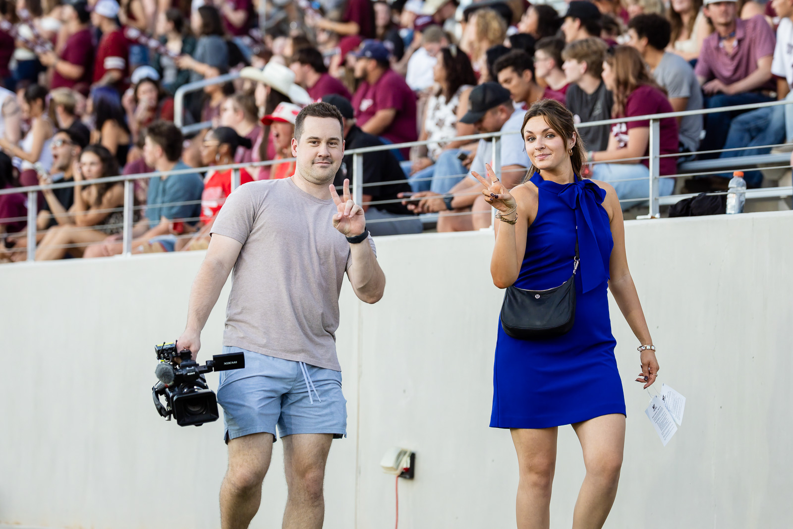 western-colorado-vs-wtamu-jpegJoe-pps-37 | Press Pass Sports Two people walk along a stadium track: a man with a video camera and a woman in a blue dress posing with peace signs amid a crowded audience.
