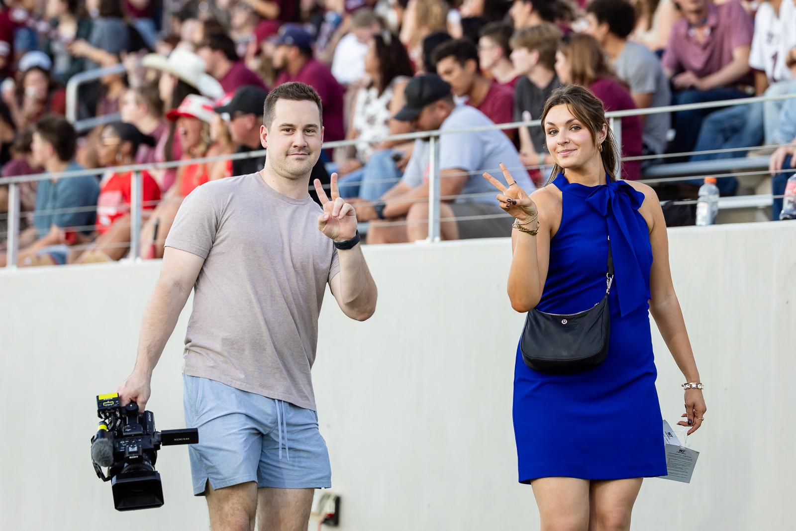 western-colorado-vs-wtamu-jpegJoe-pps-38 | Press Pass Sports Two adults pose with peace signs at an outdoor event, a cameraman on the left carrying a video camera and a woman in a blue dress on the right, with a large crowd in the stands behind them.