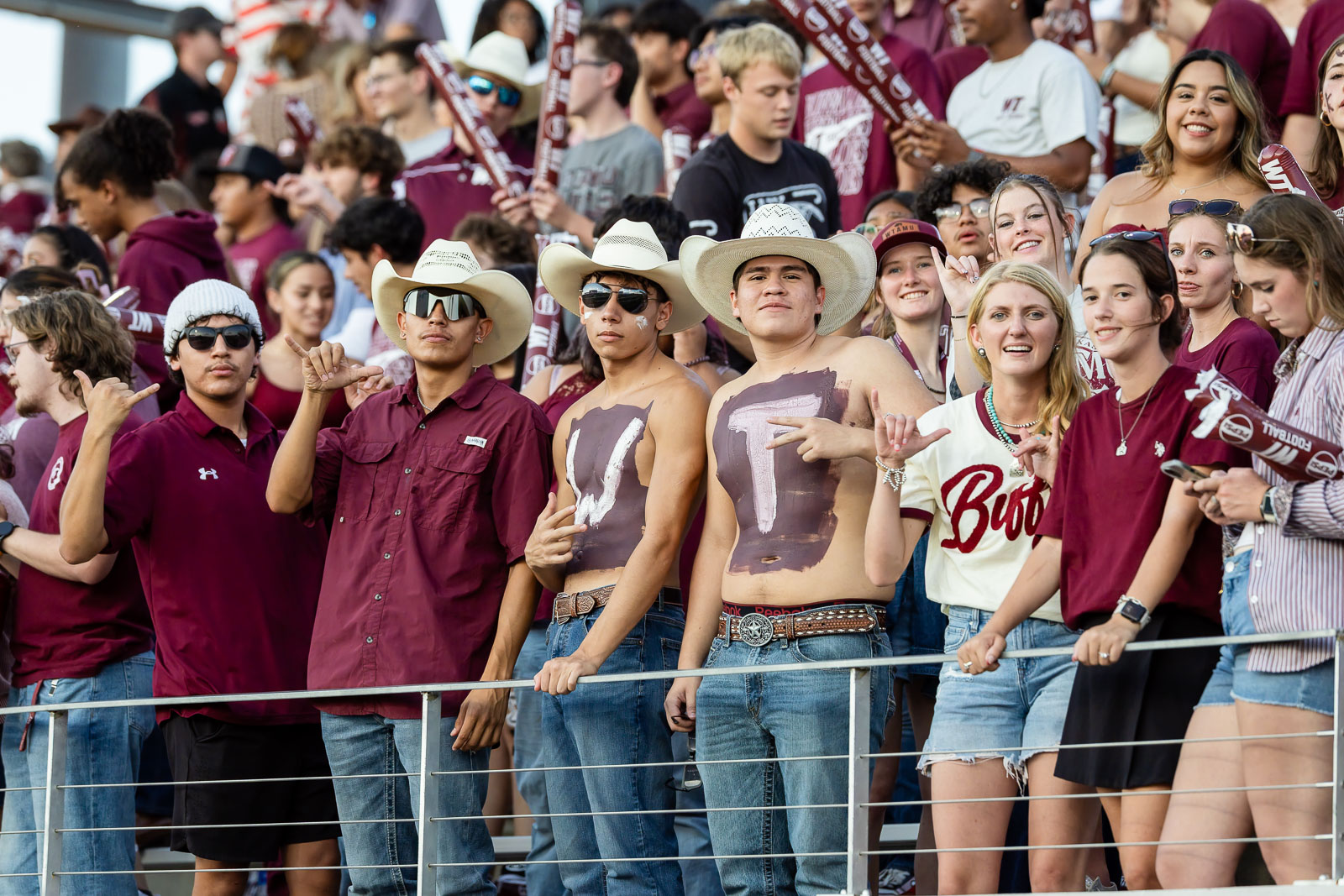 western-colorado-vs-wtamu-jpegJoe-pps-39 | Press Pass Sports Group of college fans in maroon gear cheering from a stadium rail; several wear cowboy hats, sunglasses, and body paint with large letters/logos nearby.