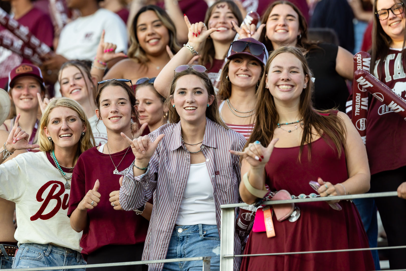 western-colorado-vs-wtamu-jpegJoe-pps-41 | Press Pass Sports Group of smiling fans in maroon clothing behind a railing at a football game, making hand gestures toward the camera.