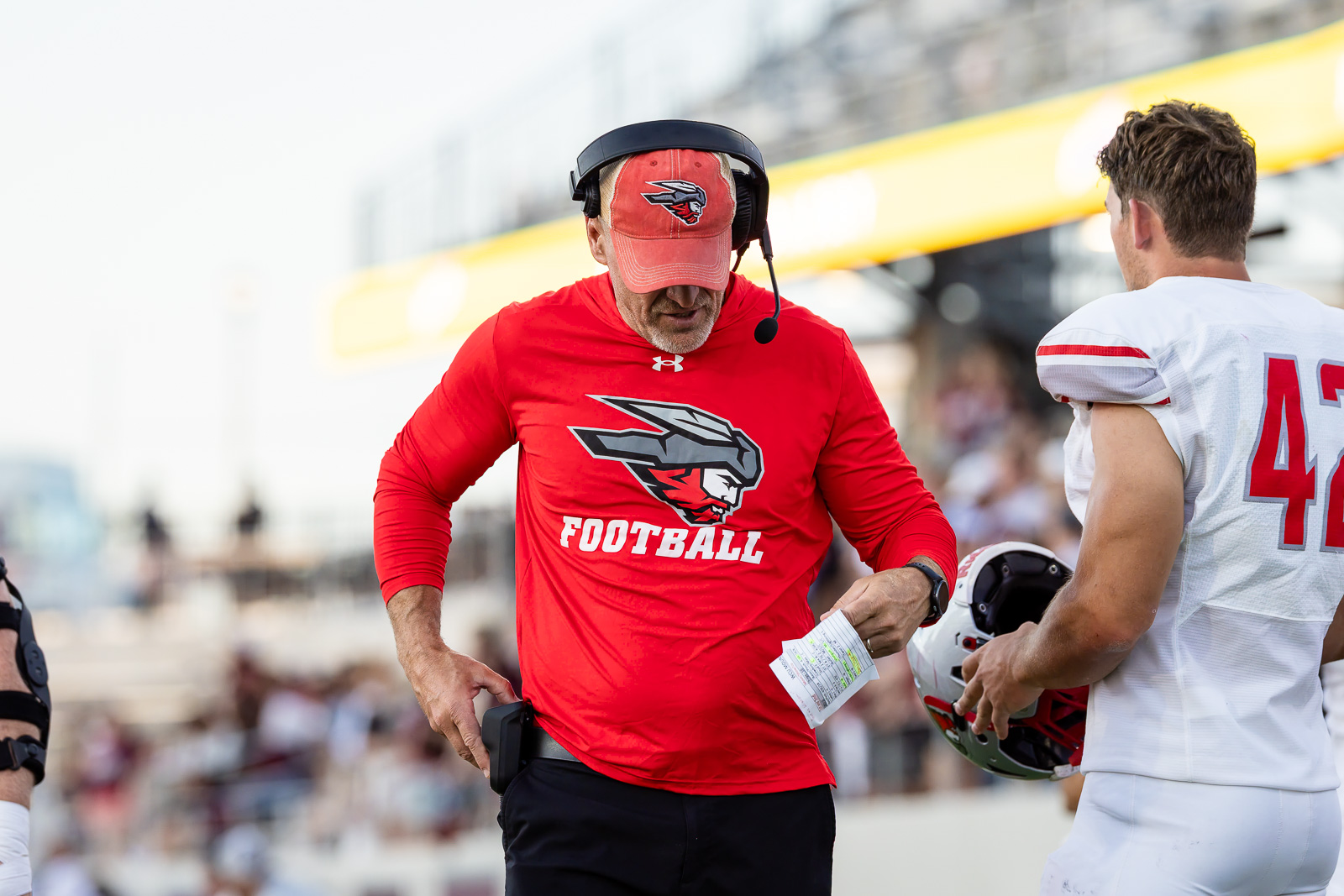 western-colorado-vs-wtamu-jpegJoe-pps-43 | Press Pass Sports Football coach in red shirt and headset talks to a player in white jersey on the sideline during a game.