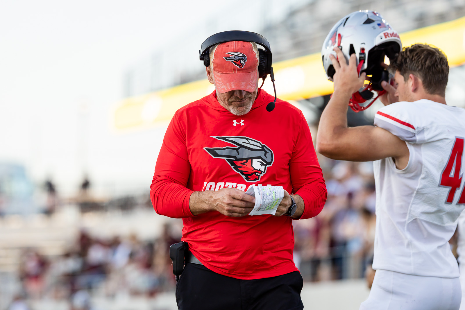 western-colorado-vs-wtamu-jpegJoe-pps-44 | Press Pass Sports Football coach in a red shirt with a headset reads a play sheet on the sideline while a player nearby holds a helmet.