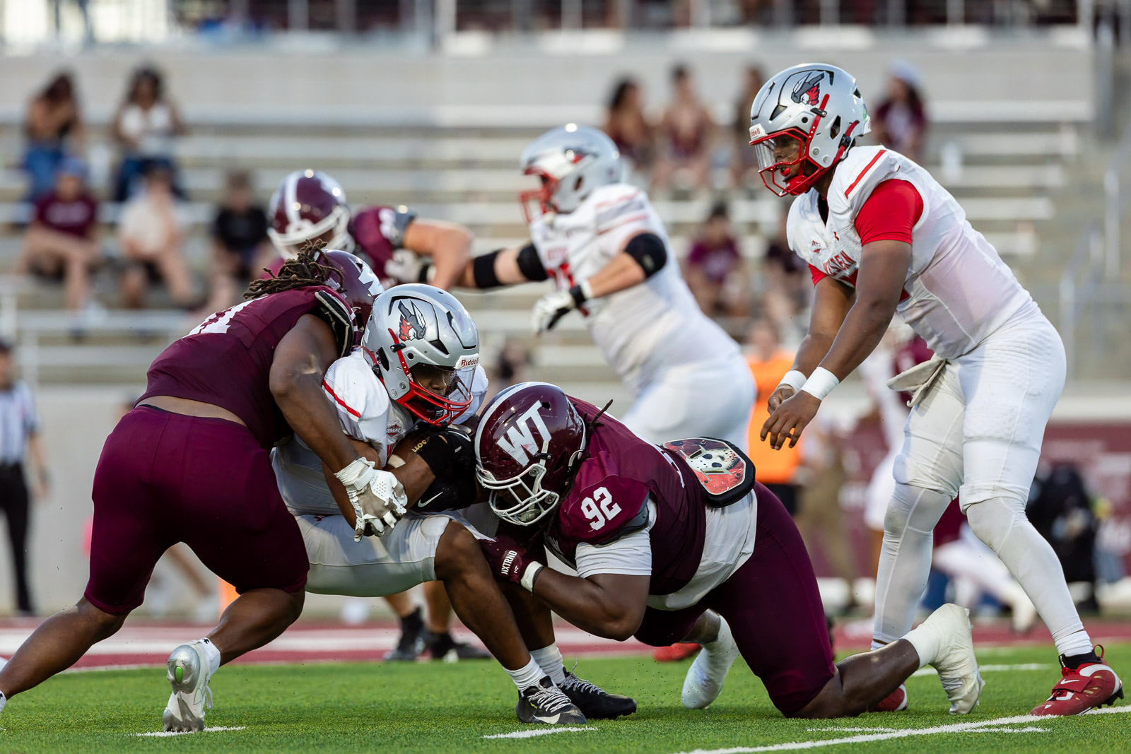 western-colorado-vs-wtamu-jpegJoe-pps-47 | Press Pass Sports College football players in maroon and white collide during a tackle on a green turf field with spectators in the stands behind them