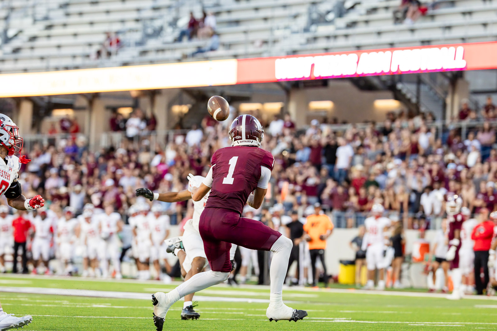 western-colorado-vs-wtamu-jpegJoe-pps-50 | Press Pass Sports Quarterback in maroon #1 runs toward a loose football in midair during a college football game, with a packed stadium in the background.