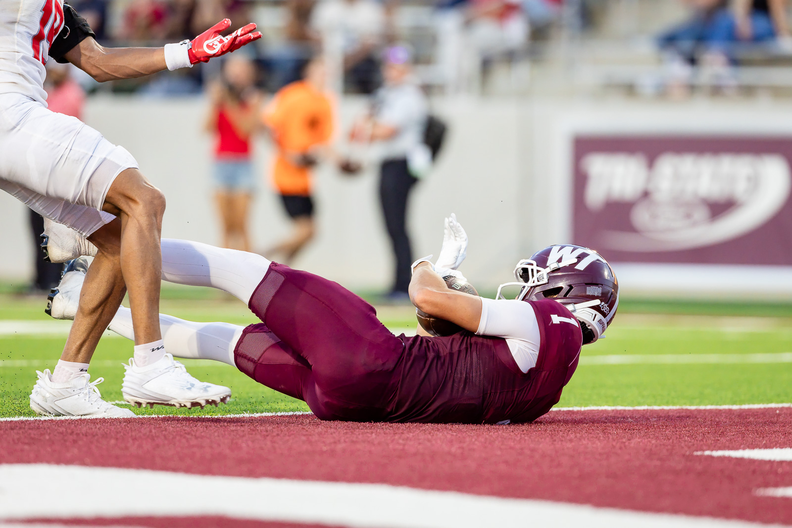 western-colorado-vs-wtamu-jpegJoe-pps-52 | Press Pass Sports Maroon football player on his side with ball, tackled on the red end zone turf as a defender stands nearby.