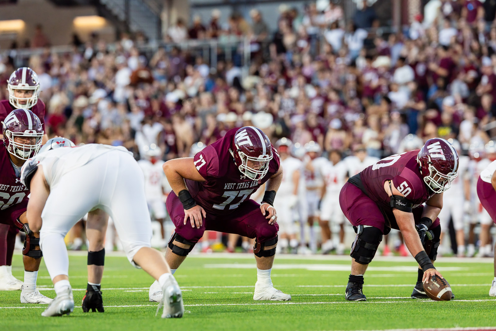 western-colorado-vs-wtamu-jpegJoe-pps-54 | Press Pass Sports American football linemen in maroon uniforms crouch at the line, ready for snap, on a green turf with a crowded stadium behind.